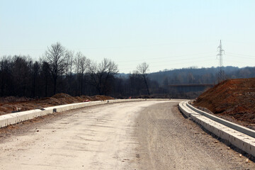 A sweeping gravel roadbed with newly installed concrete curbs curves through a construction site,...