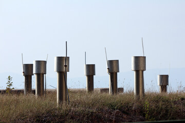 Several cylindrical metal vent pipes with caps and attached antennas are aligned in a grassy field,...