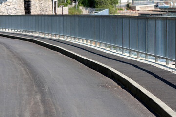 Smooth black asphalt road curves gently beside a metal safety railing, following a waterfront...