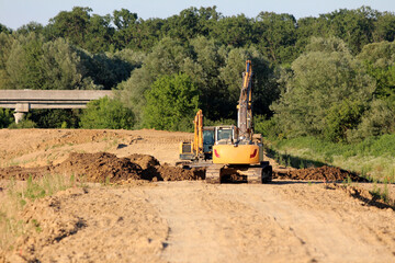 Heavy excavators dig and level earth for a new road, with forest in the background and a concrete...