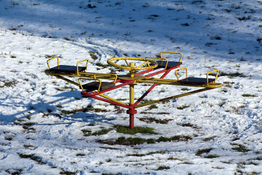 A yellow and red metal playground carousel stands on patchy snow and grass, its empty seats and shadows highlighting a cold, quiet winter day