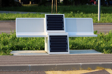 Modern white bench with integrated solar panels stands on pavement by lush grass, offering...