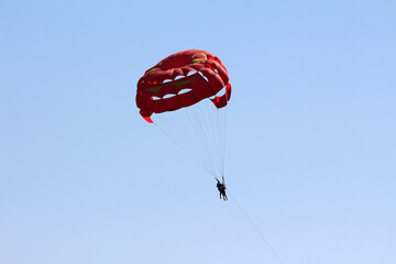 Two people are suspended beneath a bright red parasail, gliding high above with a tow line, set against a vast, cloudless sky for a thrilling aerial experience