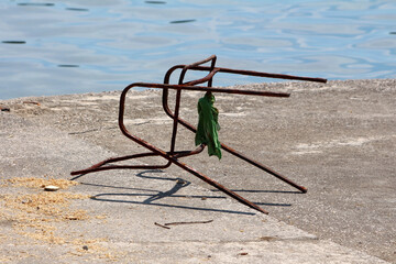 A corroded metal chair frame lies on cracked concrete near calm blue water, with a piece of green...