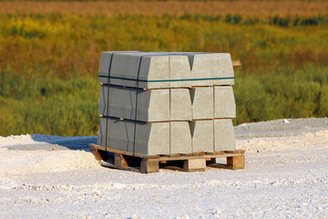 Neatly stacked concrete curbstones rest on a wooden pallet atop a white gravel surface, ready for...