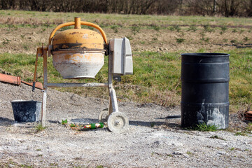 A used yellow concrete mixer stands beside a black barrel and bucket on gravel, with scattered bottles and green grass in the background