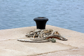 A frayed mooring rope lies coiled near a black bollard on a sunlit concrete pier, overlooking still...