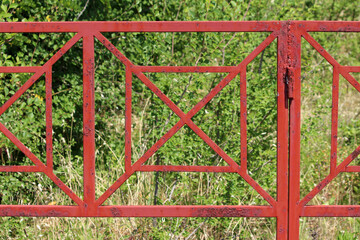 A rusty red metal gate with bold geometric shapes stands amid dense green vegetation, contrasting...