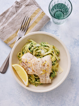 A bowl contains creamy lemon garlic pasta topped with seasoned herb cod. A slice of lemon adds freshness, complemented by a glass of water. Soft linen napkin enhances the dining presentation