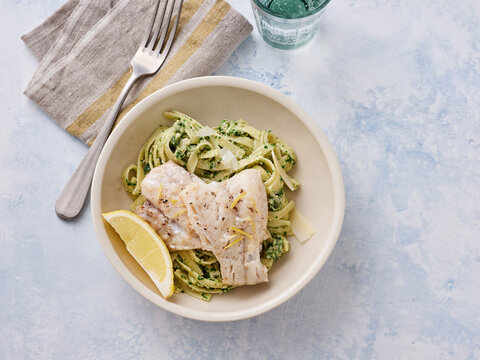 Lemon garlic pasta is served in a bowl with tender herb cod and a lemon wedge on the side. This dish combines savory and citrus flavors for a delightful dining experience