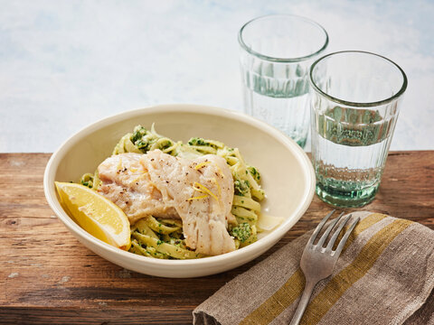 Lemon garlic pasta topped with herb cod is plated elegantly, alongside two refreshing glasses of water. The casual dining setting features a rustic wooden table