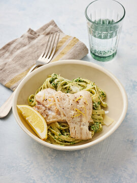 A delicious dish features lemon garlic pasta topped with herb cod, served in a simple bowl. A glass of water accompanies the meal, set on a light-colored surface