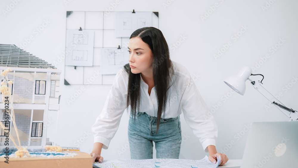Poster young beautiful caucasian architect puts blueprint on the table and compare with house model while i