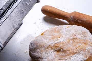 Pasta dough and dough rolling machine and rolling pin on a white table.