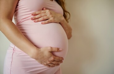 Pregnant woman dressed in pink outfit tenderly holds her baby bump. Expectant mother embraces her growing belly. Pregnancy, motherhood, anticipation of new life, prenatal care, health.