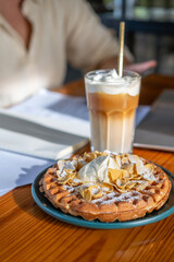 On a sunlit table, a woman balances her daily work while treating herself to a rich cheese latte and a decadent waffle topped with whipped cream, perfect for a midday break