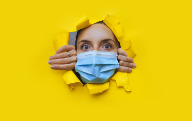 Surprised young woman in a blue medical mask looks through a torn hole in yellow paper. The concept...