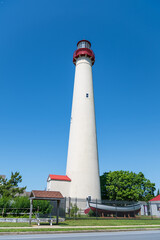 The Cape May lighthouse with a white tower and a red top stands prominently against a clear blue sky. The surrounding area features a black fence, buildings,  and a boat.