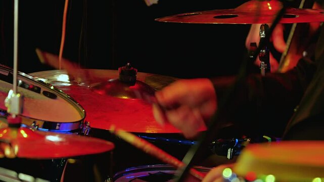The drummer is at work in close-up, his hands with sticks hitting the cymbals, and the bright light of the stage reflects off the instruments. Jazz or rock concert