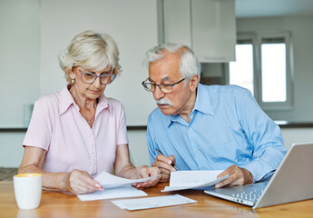 Senior couple checking bills and calculating expenses using a laptop at home, debt taxes concept,...