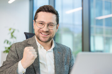 Portrait of a young successful businessman sitting in the office at a table in front of a laptop,...