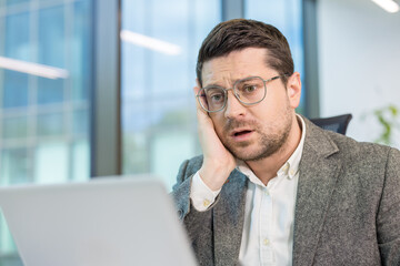 Upset young male office worker sitting at his workplace and looking disappointedly at the laptop...