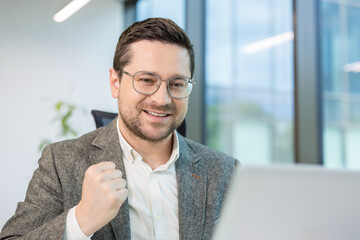 Close-up of a young smiling man sitting in the office and rejoicing in success, showing a victory...