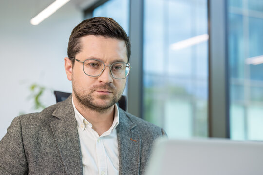 A close-up photo of a young man working in an office at a laptop, looking at the screen with concentration and thoughtfulness
