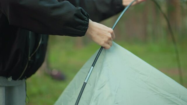 close up of woman in black jacket struggling to remove tent hook from flexible tent pole while another person hand assists partially visible in forest environment surrounded by green grass and trees