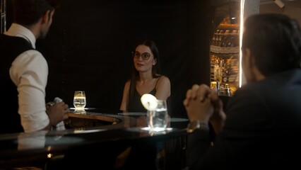 Focusing on beautiful woman guest sitting in special drink gin fizz at luxurious counter bar at vibrant nightlife party atmosphere. Smart bartender wiping glass while talking with customer. Vinosity.