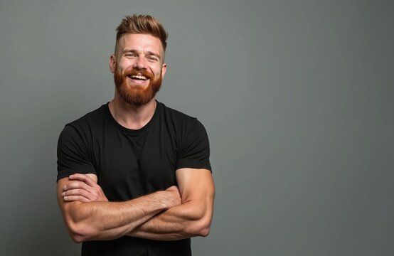Portrait of smiling muscular 30-year-old man with ginger hair and beard standing against light gray background. Confident male in black t-shirt looks cheerful with arms crossed. Healthy handsome guy.