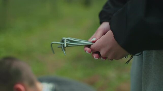 woman gives tent peg to crouching partner holding sturdy wood stick ready to hammer into soft forest ground covered with grass moss and pine needles securing tent stake during couple camping