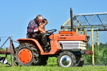 Grandfather and grandson riding a small orange tractor together in the countryside. The boy happily...