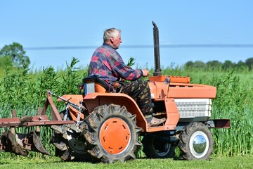 Senior farmer on compact orange tractor driving to cultivate crops in lush green field. Summer...