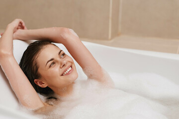 Obraz premium Relaxed young woman enjoying a bubble bath, smiling with arms raised, surrounded by soft foam and warm tones. Self-care and wellness concept.