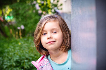 Playful preschooler peeks around pillar with mischievous gaze. Blonde bob, gray-blue eyes, light blue top. Soft summer greenery bokeh. Childhood curiosity close-up