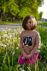 Child in dandelion field holding fluffy bloom. Dark blonde hair, pink pants, sparkling top. Intense gaze, soft backlight, nature wonder