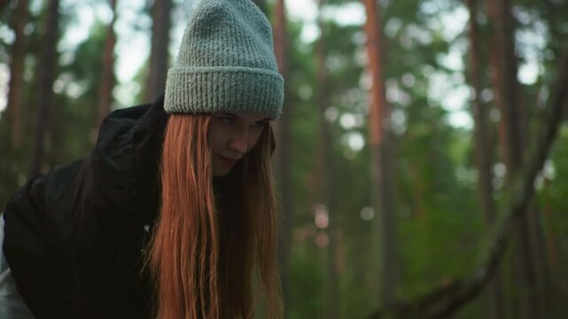 Woman in knitted cap and black jacket looks up with focused yet playful expression while engaged in outdoor task in forest, surrounded by tall trees and natural daylight filtering through leaves