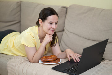 European woman lies on sofa with a laptop and a pastry, enjoying a relaxed moment at home in casual clothing, looking at the screen with a smile