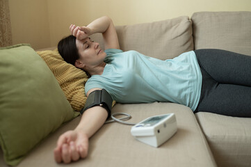 woman lies on couch with arm extended, measuring blood pressure at home, showing tired or unwell expression