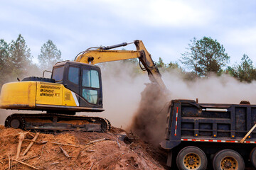Excavator moves dirt, debris into dump truck amid dust cloud on construction site surrounded by trees.