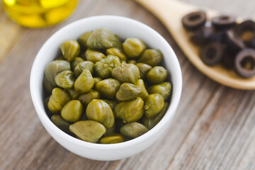 Pickled capers in small bowl with black olive slices and olive oil in the back, the ingredients for...