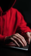 A hacker in a red hoodie types on a laptop in a dimly lit room, focusing on cybersecurity.