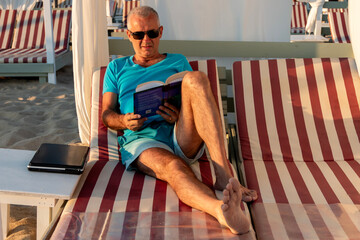 Mature man reading a book and relaxing on a daybed at the beach. Travel, weekend, relax and...