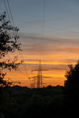 Fototapeta premium A tall electricity transmission tower and power lines cast silhouettes against a dramatic dusk sky streaked with colorful clouds over a lush treeline