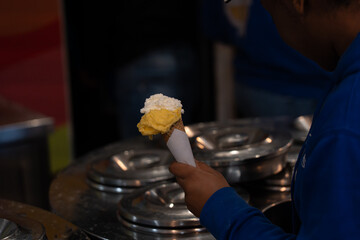 Hands clad in a blue hoodie scoop soft-serve ice cream into a golden waffle cone at a busy parlour...