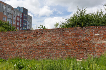 Old red brick wall with weathered texture and overgrown grass in front of apartment buildings on a...