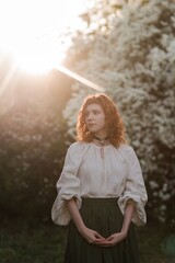 Young pretty caucasian woman in romantic outfit walking in blooming spring garden. Artistic outdoor photo of beautiful red-haired girl posing in fragrant lilac blossom.