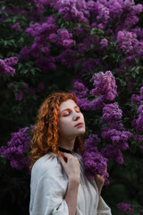 Young pretty caucasian woman in romantic outfit walking in blooming spring garden. Artistic outdoor photo of beautiful red-haired girl posing in fragrant lilac blossom.