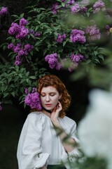 Young pretty caucasian woman in romantic outfit walking in blooming spring garden. Artistic outdoor photo of beautiful red-haired girl posing in fragrant lilac blossom.